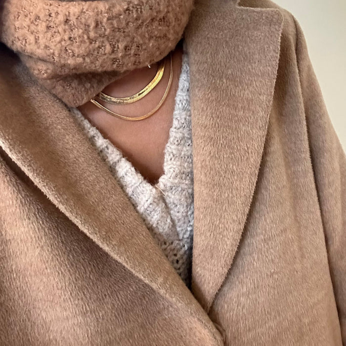 A close-up of a person in a light brown coat, chunky knit scarf, and sweater, accessorized with the Hailey Duo snake chain necklace by LLUME Jewelry. The photo highlights the outfit’s layered textures and elegant jewelry detail.