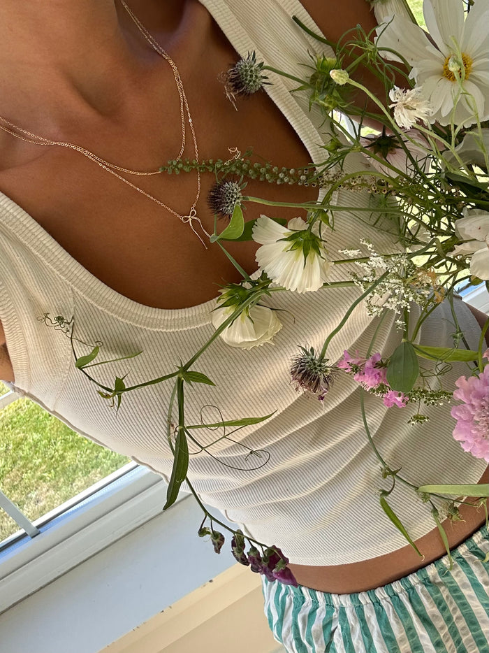 Wearing a white ribbed tank and striped pants, a person holds wildflowers close, layered in shining silver necklaces including the LLUME Jewelry Bow Daily Set, as they stand by a sunlit window.