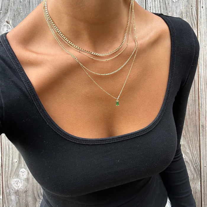 Wearing the LLUME Jewelry Kismet Solstice Set, featuring a gold layered necklace with an emerald green pendant, and a black scoop-neck top, a person stands before weathered wooden boards.
