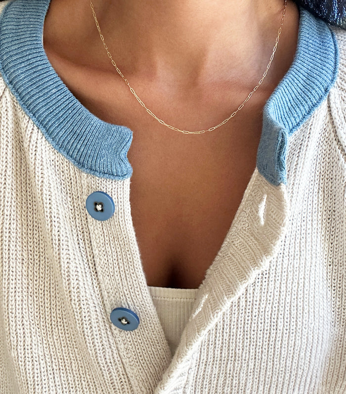 A close-up of a person wearing the LLUME Jewelry Adelene Paperclip Chain, styled with a cream knit cardigan with blue trim and buttons over a white top. The image highlights the neckline and upper chest area.