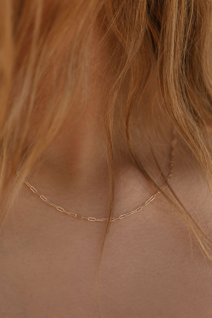 Close-up of a woman's neck with wavy, light brown hair wearing the Adelene Paperclip Chain by LLUME Jewelry—an 18k gold plated necklace with small, elongated links.