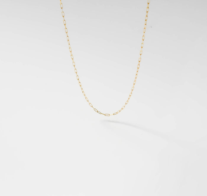 The Adelene Paperclip Chain by LLUME Jewelry features small, oval gold-plated links. Shown against a white background, the adjustable necklace forms a gentle curve and casts a subtle shadow below.