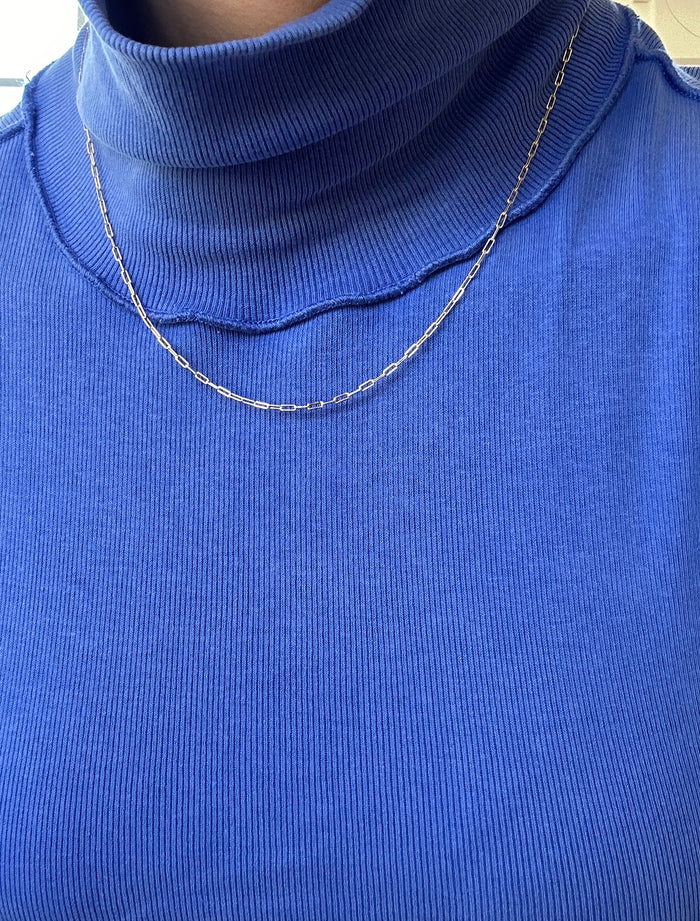 Someone wears a bright blue ribbed turtleneck top paired with the Adelene Paperclip Chain by LLUME Jewelry, a thin and delicate gold-plated necklace. The photo highlights the chest and neckline.
