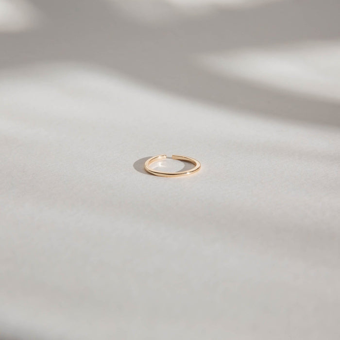 An LLUME Jewelry Adjustable Stacking Ring in 14k gold-fill rests on a smooth, light-toned surface, with gentle, blurred shadows in the background.