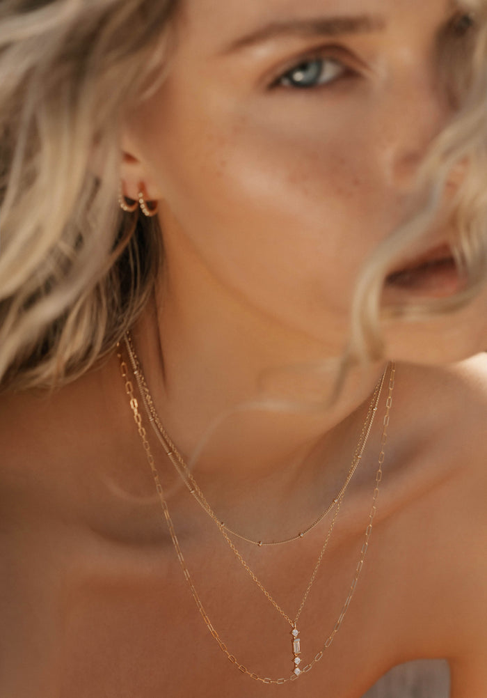 Close-up of a woman with wavy blonde hair wearing the LLUME Jewelry Adorn Set, featuring layered necklaces and small gold hoops. The soft focus highlights her delicate jewelry and natural skin.