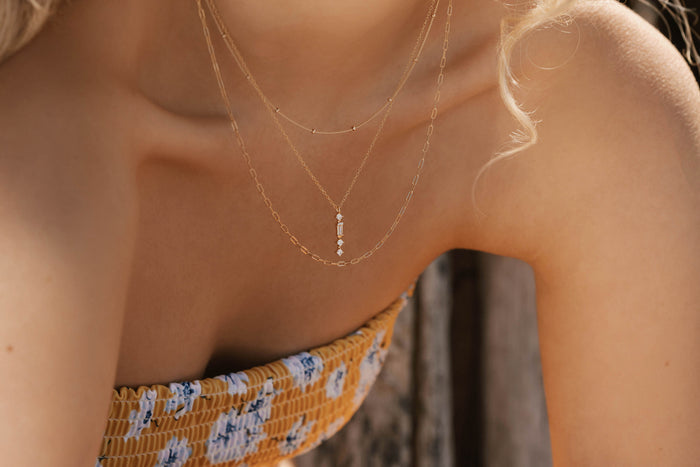 Close-up of a woman wearing the Adorn Set by LLUME Jewelry—a versatile set of layered gold necklaces with small pendants. She pairs it with a yellow and white floral strapless top, her blonde hair partially visible.