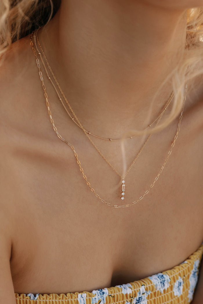 A woman in a yellow floral top wears the Adorn Set by LLUME Jewelry—three delicate layered gold necklaces, one featuring a small vertical pendant with tiny beads—seen from the shoulders up.