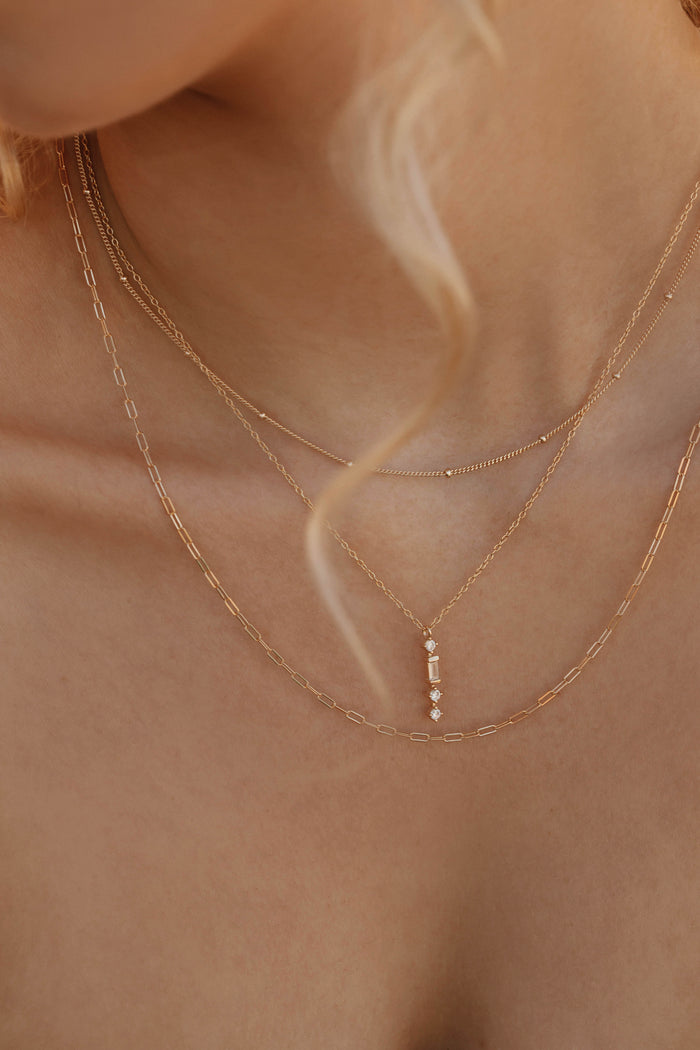 Close-up of a woman's neck and collarbone wearing the Adorn Set by LLUME Jewelry—layered delicate gold necklaces, including a pendant with small vertical gemstones; a soft curl of blonde hair is also visible.