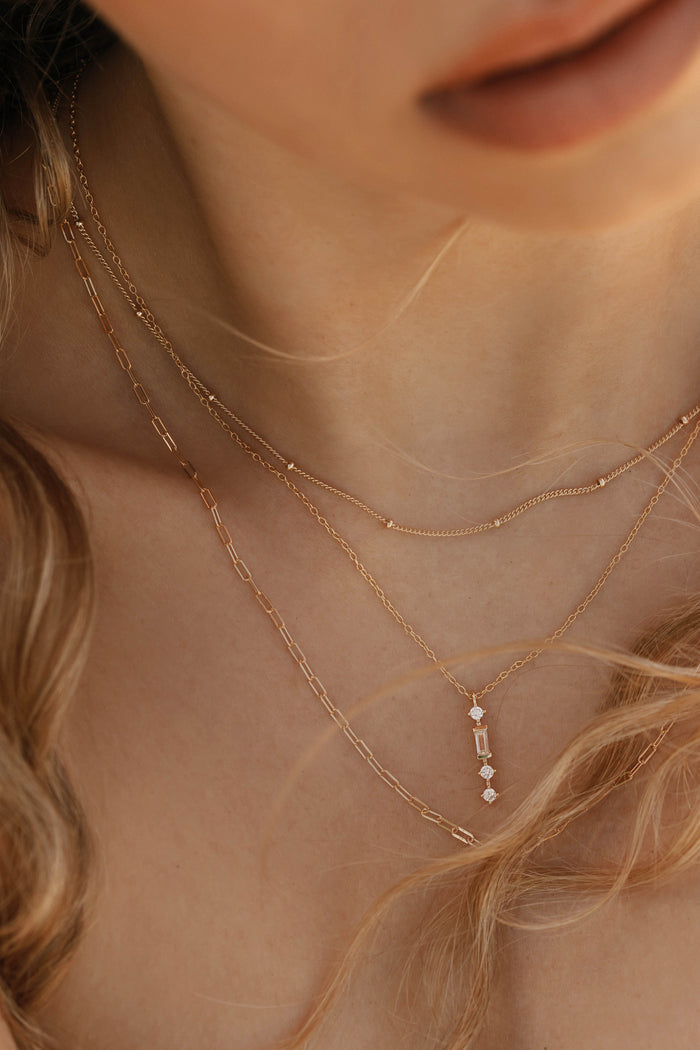 A close-up shows a woman’s neck adorned with LLUME Jewelry’s Adorn Set: three delicate gold chains—one with rectangular gemstones—framed by wavy blonde hair, highlighting this versatile necklace set.