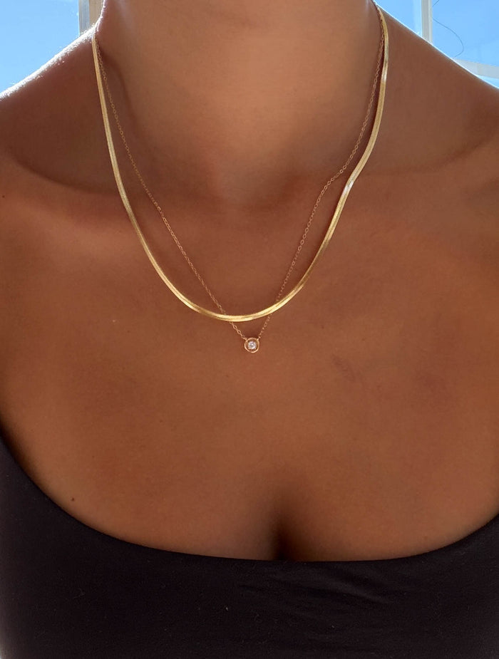 A close-up of a person’s neck and upper chest showcasing the Alexis Set by LLUME Jewelry, layered with a simple chain, against a blue sky. The person is also wearing a black strapless top for an effortless look.