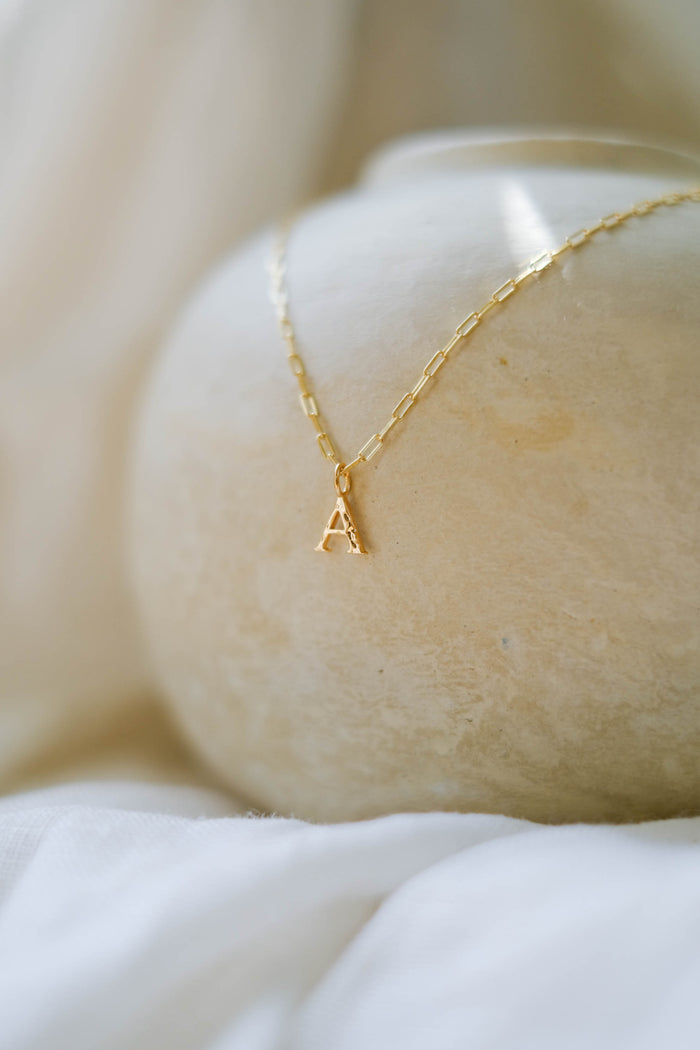 The LLUME Jewelry Alphabet Charm Necklace, a delicate gold piece featuring a customizable letter A charm, rests on a smooth light stone with soft white fabric in the background.