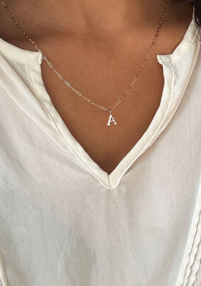 A person wears the LLUME Jewelry Alphabet Charm Necklace with a letter A charm, styled with a cream V-neck top.