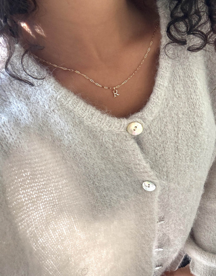 A person wears a soft, light gray fuzzy cardigan with large white buttons and an LLUME Jewelry Alphabet Charm Necklace featuring an R pendant; their curly hair is partially visible.