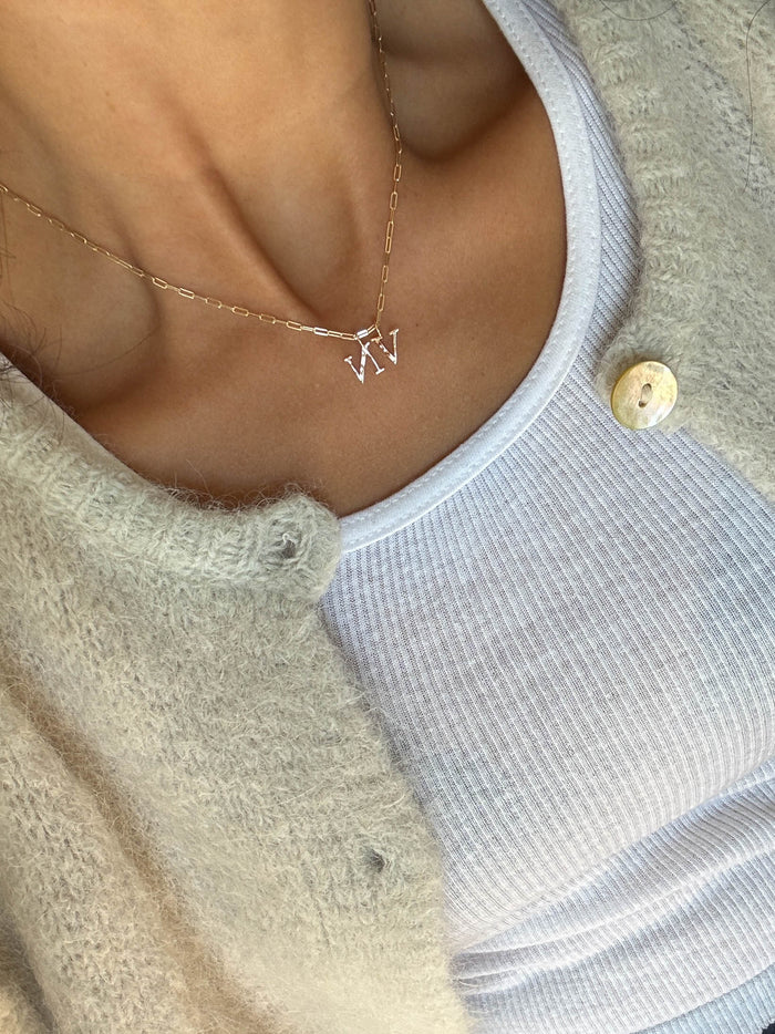A close-up of a person wearing the LLUME Jewelry Alphabet Charm Necklace with two letters, paired with a white ribbed tank top and a beige textured cardigan featuring a large button.