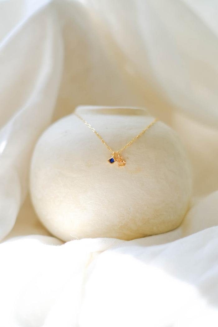 The LLUME Jewelry Alphabet Charm Necklace, a delicate gold customizable piece with a blue gemstone pendant, is draped over a round textured white vase and surrounded by soft white fabric folds.