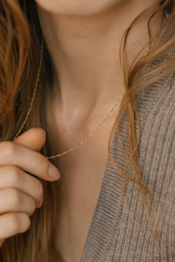 A woman holds the elegant LLUME Jewelry Angel Twist Necklace near her neck, wearing a taupe ribbed sweater with her long brown hair partially visible.