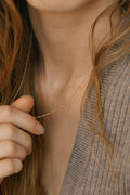 A woman holds the elegant LLUME Jewelry Angel Twist Necklace near her neck, wearing a taupe ribbed sweater with her long brown hair partially visible.