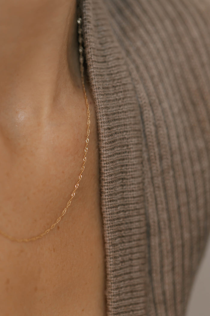 A close-up of a person's neck wearing the LLUME Jewelry Angel Twist Necklace—a delicate, gold-plated piece—paired with a ribbed brown knit top. The focus highlights the necklace's details and the fabric's texture.