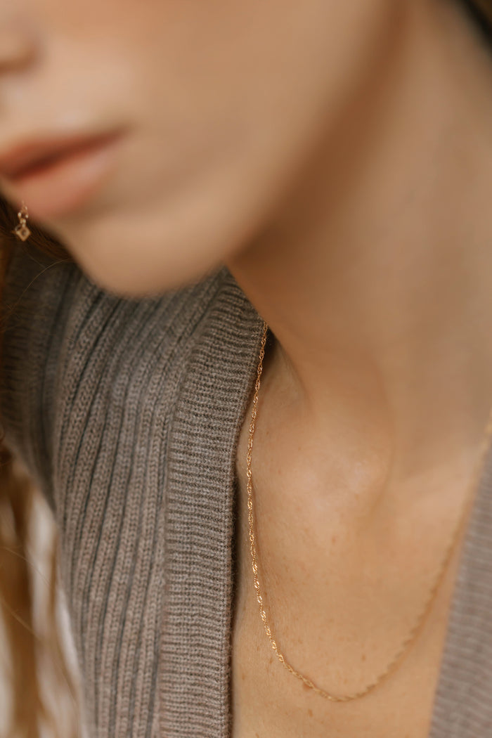 Close-up of a woman in a gray ribbed sweater wearing the Angel Twist Necklace by LLUME Jewelry, a delicate gold-plated piece, with part of her face out of focus and lips slightly visible.