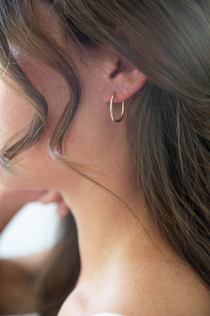 Close-up of a woman with long brown hair wearing LLUME Jewelry's Athena Oval Hoops; a loose curl frames her cheek while the softly blurred background highlights the vintage-inspired gold earrings.