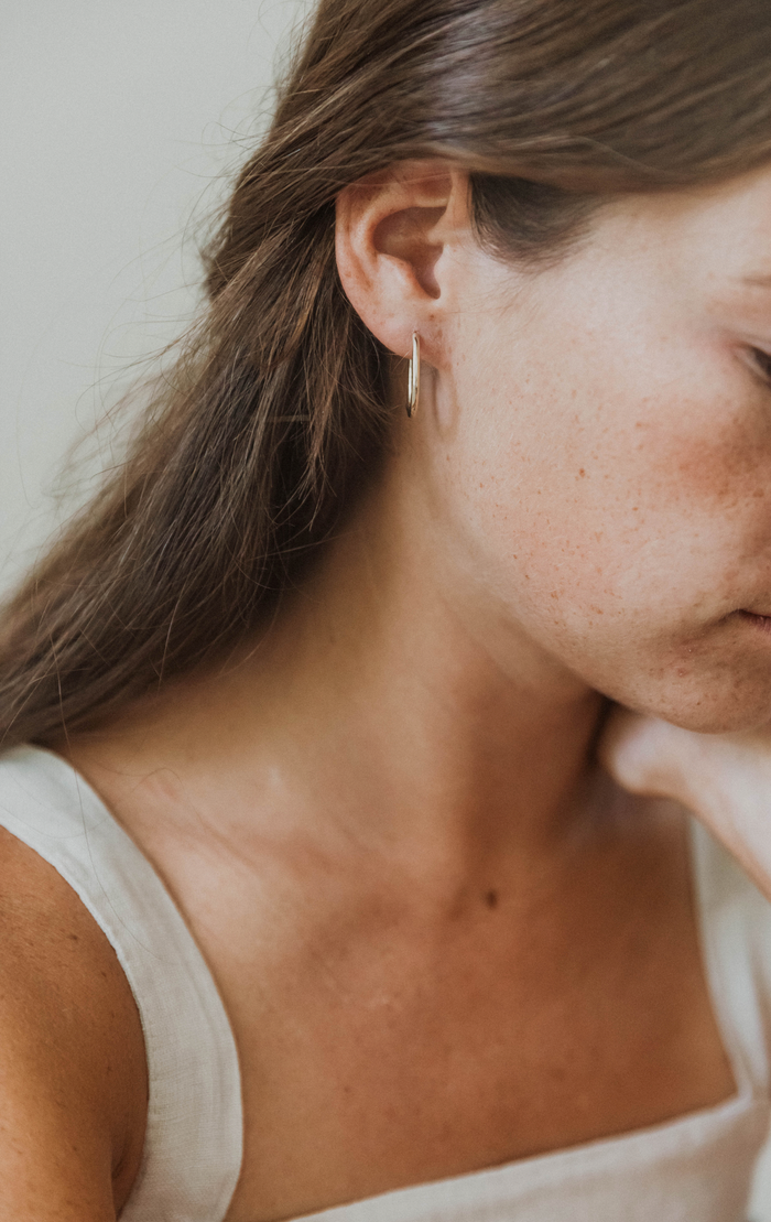 A close-up of a woman with long brown hair wearing a white sleeveless top and LLUME Jewelry's Athena Oval Hoops. Her head is slightly tilted down, and her face is partially out of the frame.