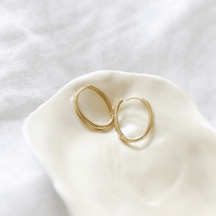 The Athena Oval Hoops by LLUME Jewelry, two vintage-inspired gold earrings, are displayed on a cream ceramic dish with a soft white fabric backdrop.