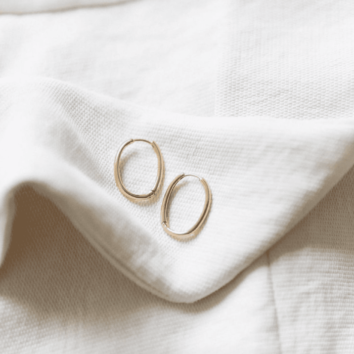 The Athena Oval Hoops by LLUME Jewelry, featuring 18k gold plating and secure leverback closures, rest elegantly on soft, white textured fabric.
