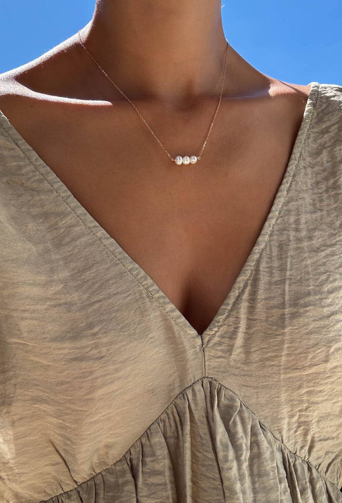 A close-up of a person wearing a beige, textured V-neck dress and the Avery Pearl Necklace by LLUME Jewelry, featuring three small freshwater pearls, set against a clear blue sky.