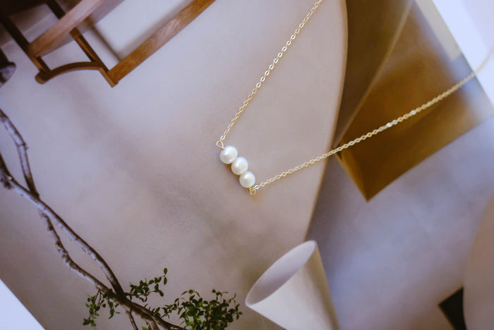 The Avery Pearl Necklace by LLUME Jewelry, a delicate gold piece with three freshwater pearls, is showcased on an open magazine with modern neutral-toned interior decor in the background.