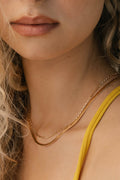Close-up of a woman’s wavy blonde hair, natural lips, and neck adorned with LLUME Jewelry’s Beverly Set—a collection of timeless layered chains. She exudes understated elegance in a yellow thin-strap top.
