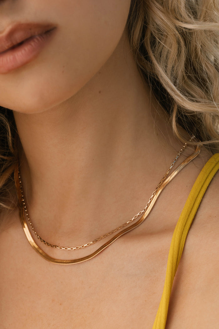 A woman with light skin and wavy blonde hair wears LLUME Jewelry’s Beverly Set layered necklaces and a yellow top. The timeless chains catch the soft, natural light, showcasing understated elegance.