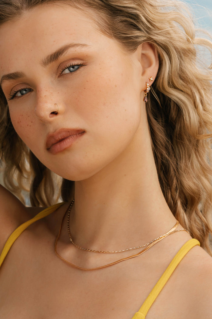 A young woman with wavy blonde hair wears the Beverly Set by LLUME Jewelry—timeless gold chains and layered necklaces. She has a nose ring, light freckles, and understated elegance in a yellow top against a soft, blurred background.