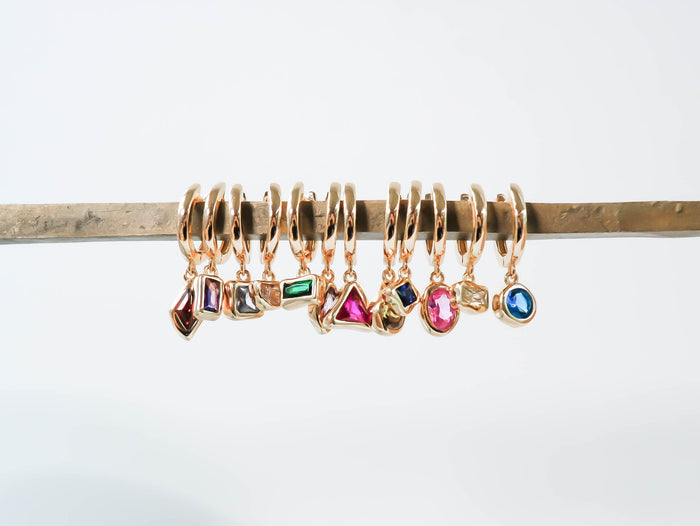 LLUME Jewelry’s Birthstone Earrings feature nine gold-plated styles, each with a unique colorful gemstone charm, displayed on a brass rod—perfect as personalized jewelry or for celebrating each birth month.