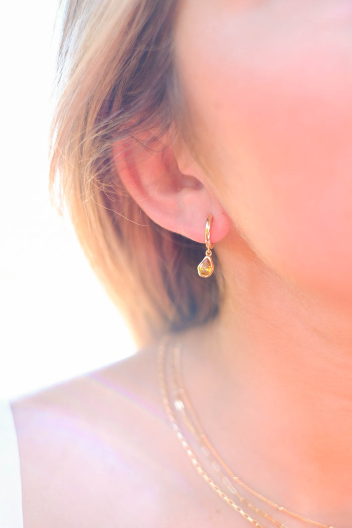 Close-up of a woman's ear wearing LLUME Jewelry's Birthstone Earrings. She also wears layered gold necklaces. The bright, softly focused background highlights the elegant gold-plated earrings.