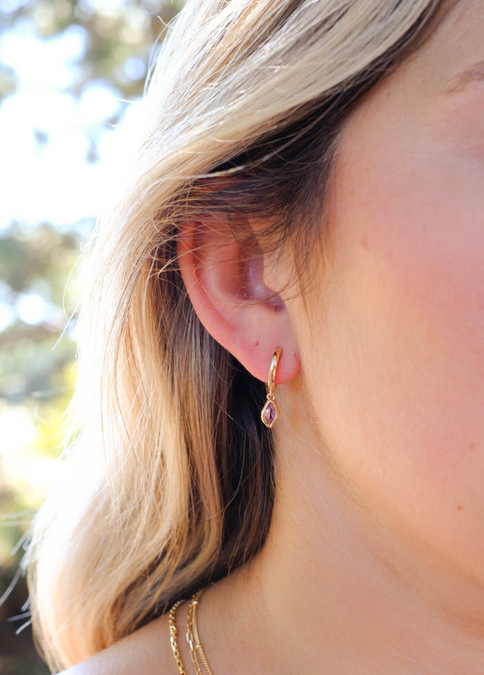 A close-up showcases a woman's ear wearing LLUME Jewelry Birthstone Earrings, gold plated with a dangling purple stone. Her blonde hair is partly shown, while natural light creates a softly blurred background glow.