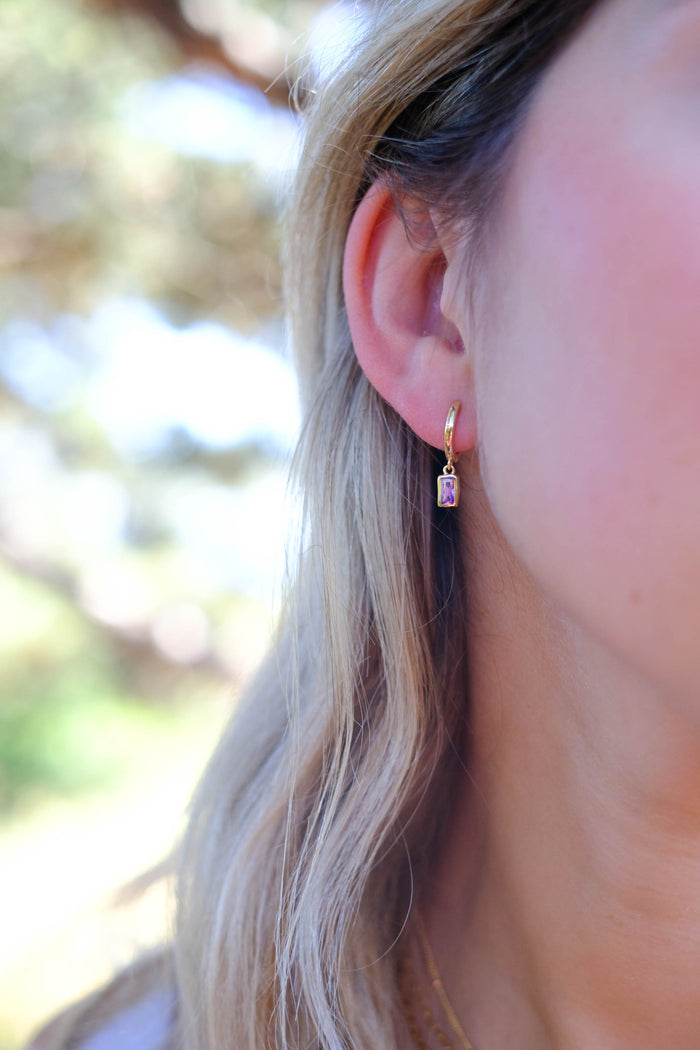 A woman with light blonde hair wears LLUME Jewelry’s Birthstone Earrings—a small gold plated design with a rectangular pendant. The softly blurred background highlights their delicate, personalized style.