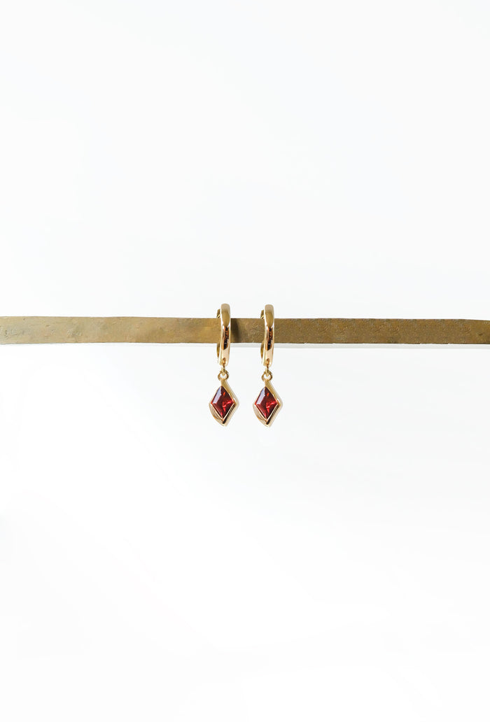 The LLUME Jewelry Birthstone Earrings feature small, square red birthstone pendants on gold-plated hooks, elegantly displayed on a horizontal brass rod against a plain white background.