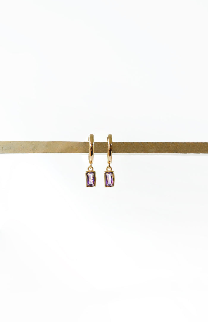 LLUME Jewelry's Birthstone Earrings feature gold plated hoops with small rectangular purple gemstone pendants, displayed on a brass rod against a white background—perfect for personalized style.
