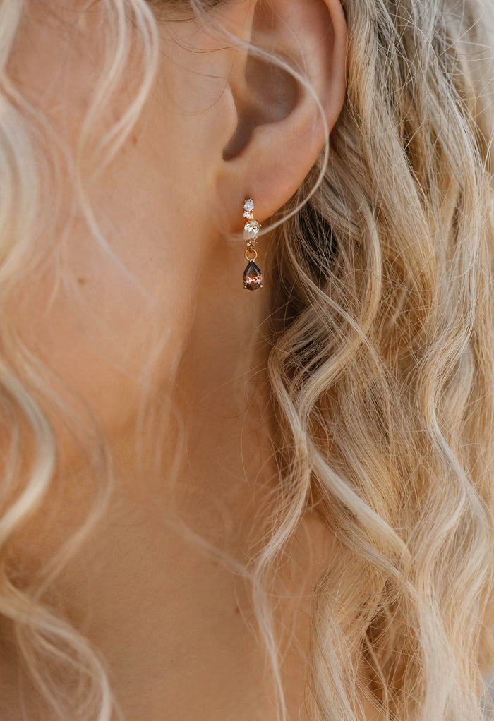 A close-up shows a woman's ear with long, wavy blonde hair, wearing LLUME Jewelry's Blair Earrings—elegant accessories with a small clear stone and a brown teardrop-shaped gemstone in a unique design.