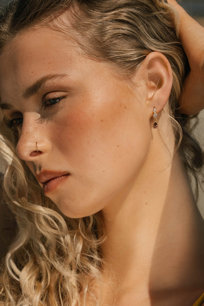 A woman with wavy blonde hair, fair skin, and freckles wears LLUME Jewelry's Blair Earrings—unique pieces that add elegance. She gazes downward, looking pensive with her hand behind her head.