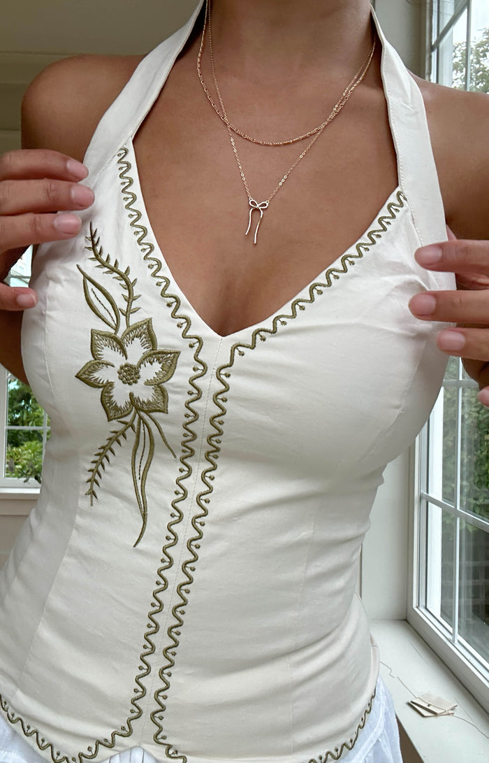 A person wearing a white halter top with floral embroidery and zigzag stitching, along with the Bow Daily Set from LLUME Jewelry, stands near a window with greenery outside.
