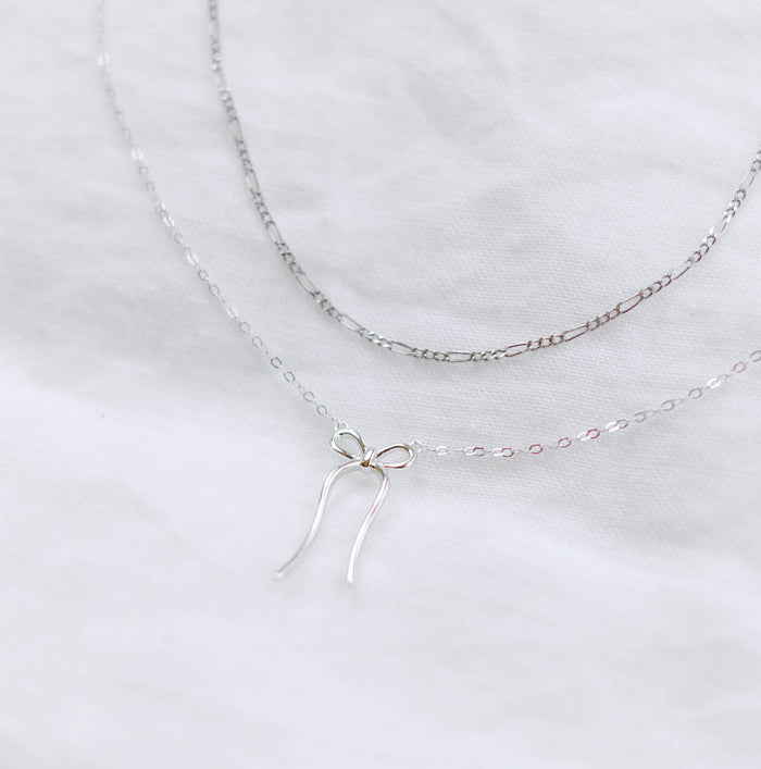 Two delicate silver necklaces from LLUME Jewelry’s Bow Daily Set rest on a white fabric—one classic chain and the other with a charming bow pendant. This elegant duo is perfect for effortless layering.