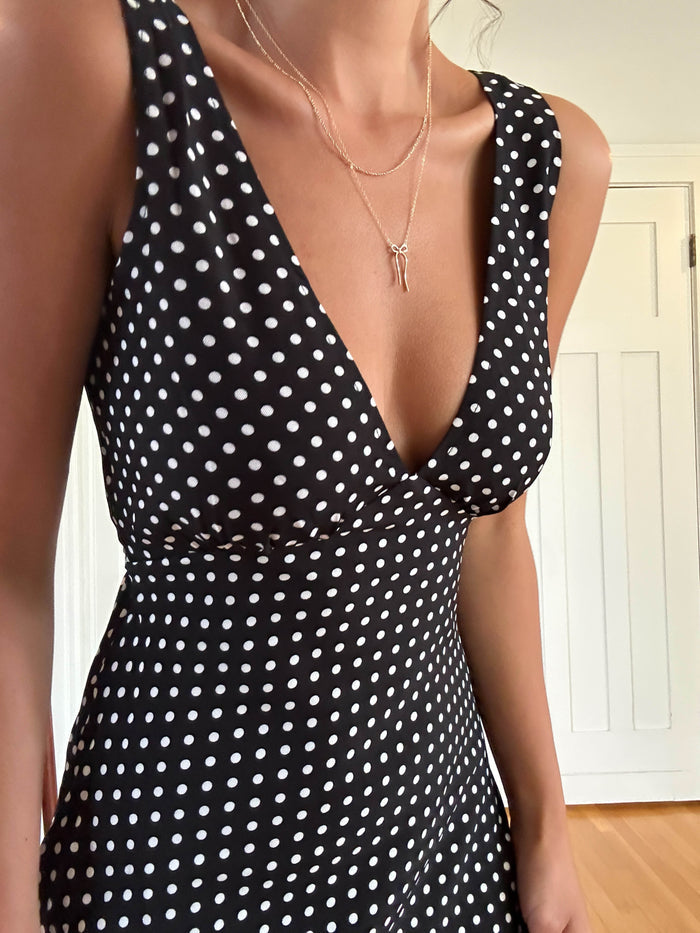 A person wearing a sleeveless black dress with white polka dots and the Bow Daily Set by LLUME Jewelry stands indoors near a white door.