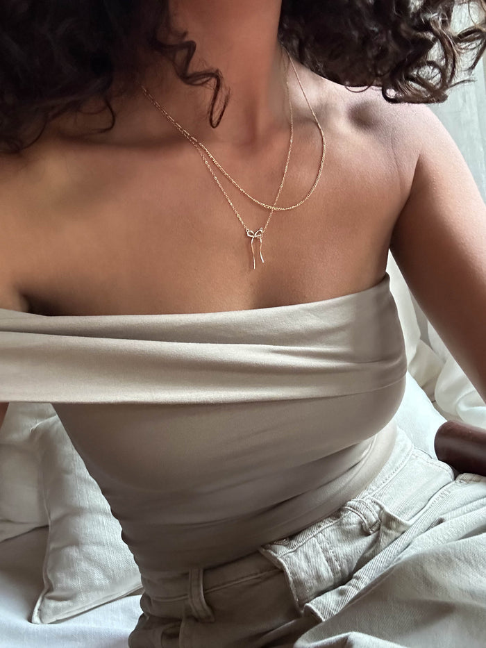 A woman with curly hair wears a cream off-shoulder top, light pants, and the LLUME Jewelry Bow Daily Set layered with a gold chain while sitting on a bed with white sheets. The image is cropped to her upper body.