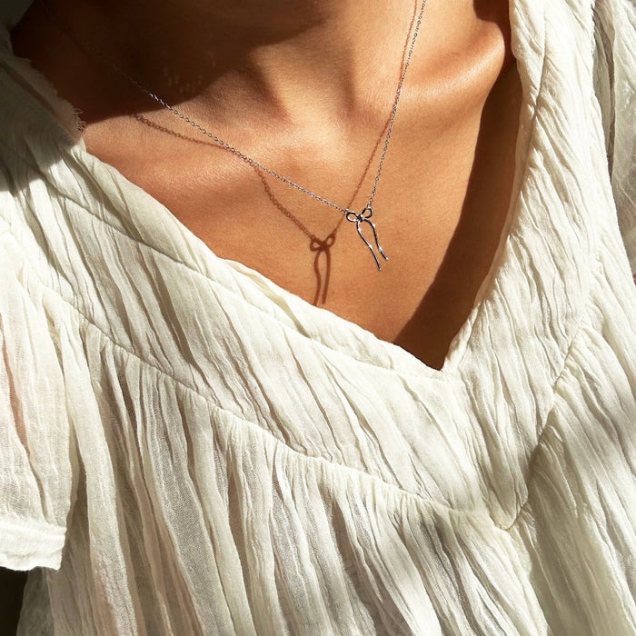 Close-up of a person wearing the LLUME Jewelry Bow Necklace (limited edition, silver), casting a delicate shadow on their chest. They are dressed in a white, textured, pleated blouse.