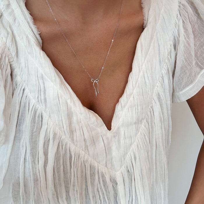A person wears a textured white V-neck blouse and LLUME Jewelry’s Bow Necklace, a limited edition gold-plated piece with a petite bow pendant. The image is cropped to highlight the neck, collarbone, and upper chest.