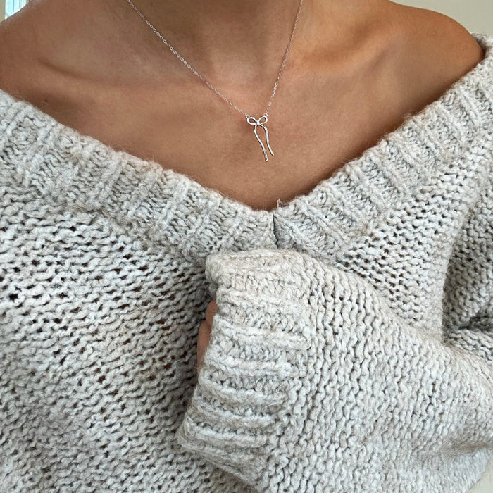A person in a chunky, light beige knitted sweater holds the collar close to their chest, wearing the LLUME Jewelry Bow Necklace—a delicate, limited edition gold plated piece.