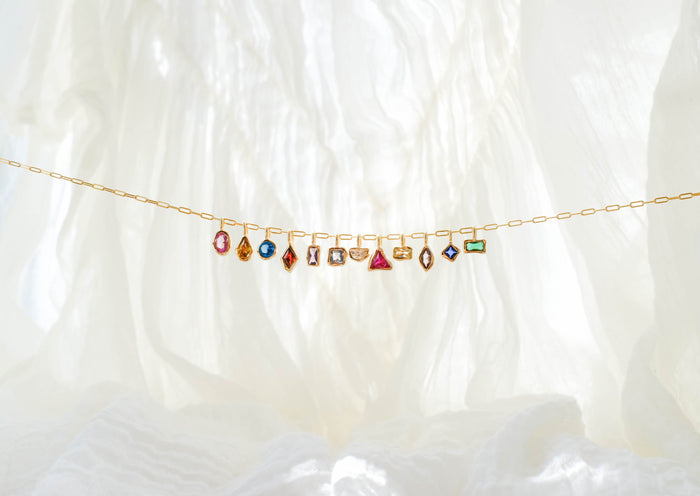 The LLUME Jewelry Build a Birthstone Necklace, featuring a delicate gold chain with customizable colorful gemstone and gold plated charms, is displayed against a softly lit white fabric background.