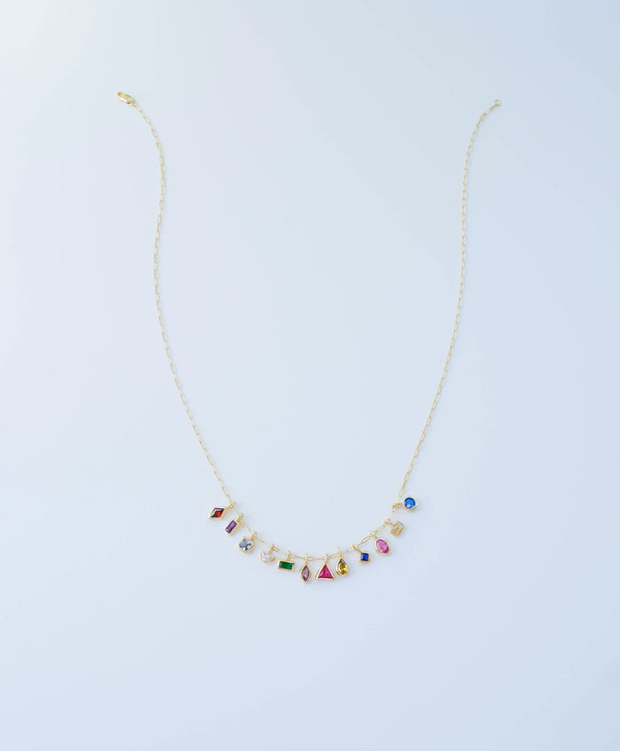 The LLUME Jewelry Build a Birthstone Necklace features gold-plated custom charms and small, colorful birthstone gems in various shapes, evenly spaced along the lower chain, displayed on a plain light background.