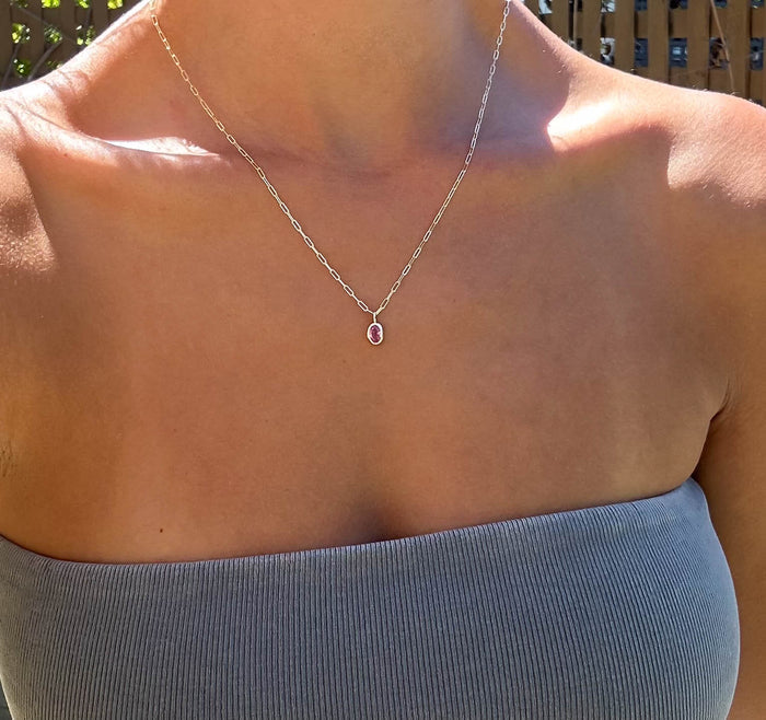 A person outdoors in sunlight wears a gray strapless top and the LLUME Jewelry Build a Birthstone Necklace—a thin silver chain with an oval pink pendant—around their neck.